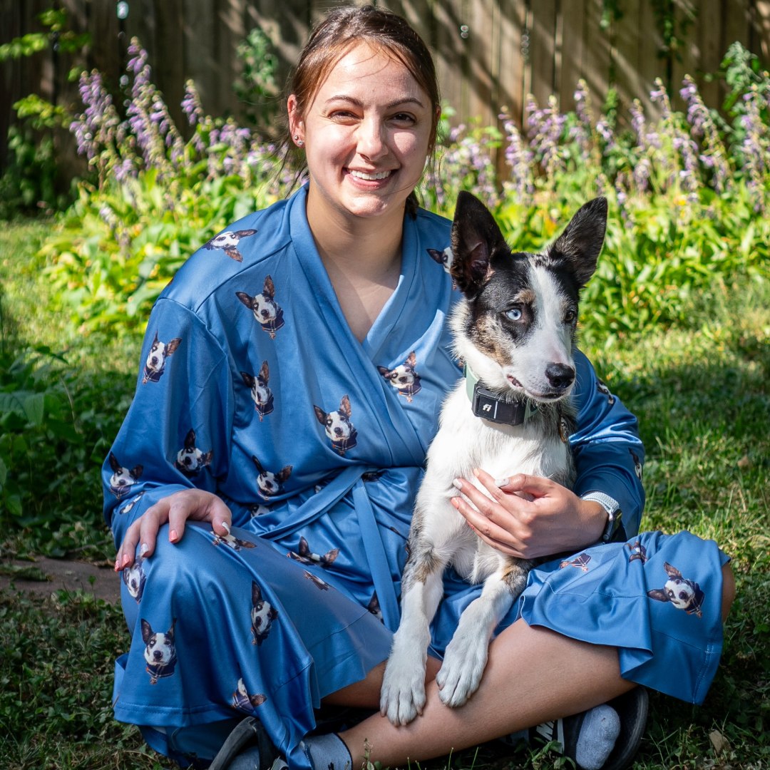 A woman sitting on the grass wearing a blue robe printed with multiple images of her dog’s face, smiling while holding a black-and-white dog with blue eyes in her lap.