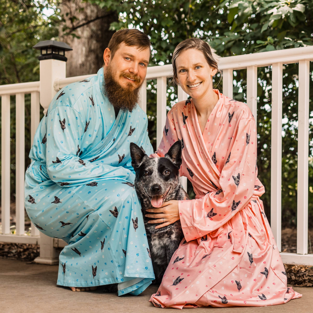 A man and woman kneel on a porch beside their black-and-white dog. They are wearing matching custom robes—his in light blue and hers in pink—both printed with the dog’s face and small paw-print designs.