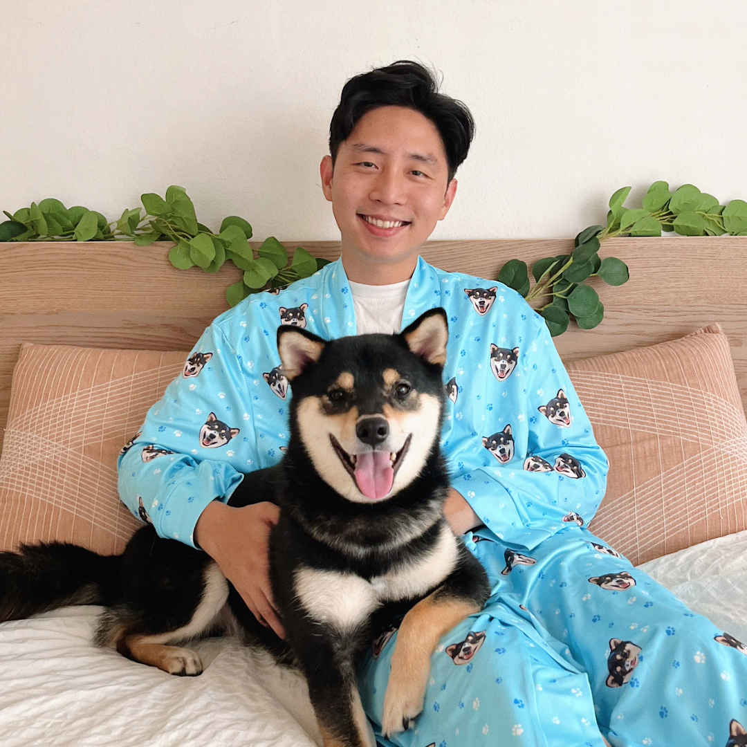 A smiling man in light blue pajamas patterned with Shiba Inu faces sits on a bed with his black and tan Shiba Inu on his lap.