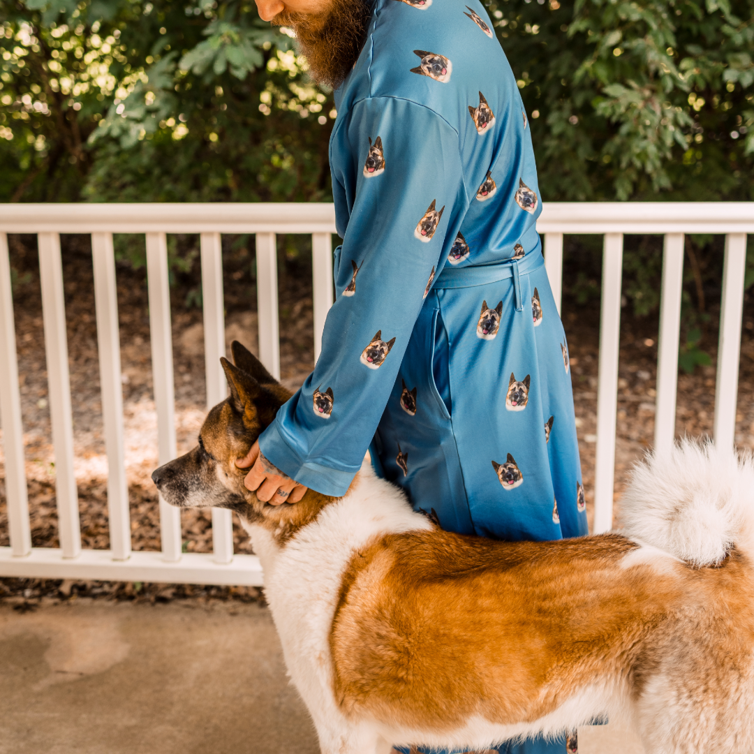 A man wearing a blue robe printed with repeated images of a dog is standing on a porch, gently holding and petting a large brown-and-white dog.