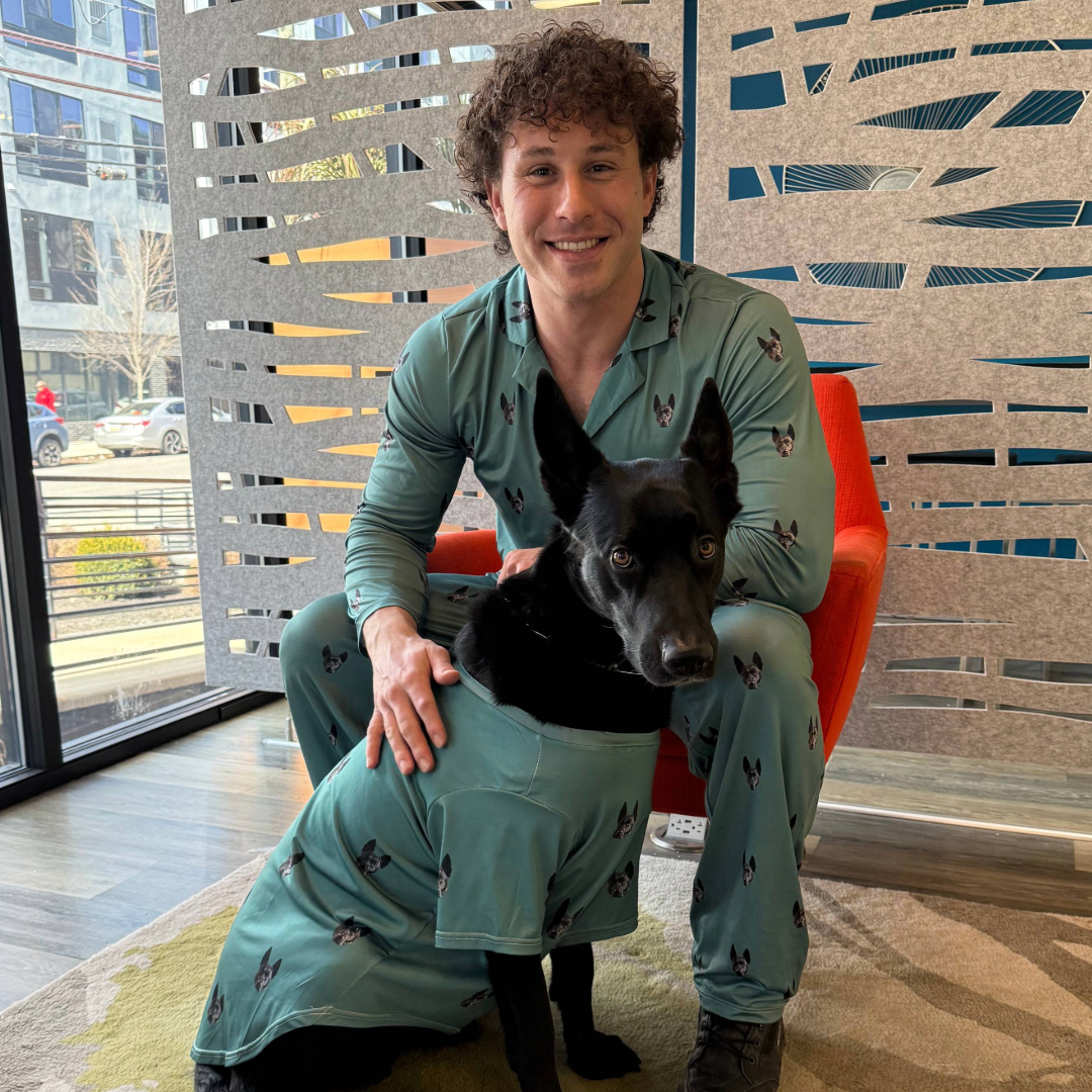 A man wearing teal pajamas printed with a black dog’s face sits in an orange chair while gently holding a black dog dressed in a matching teal shirt with the same pattern.