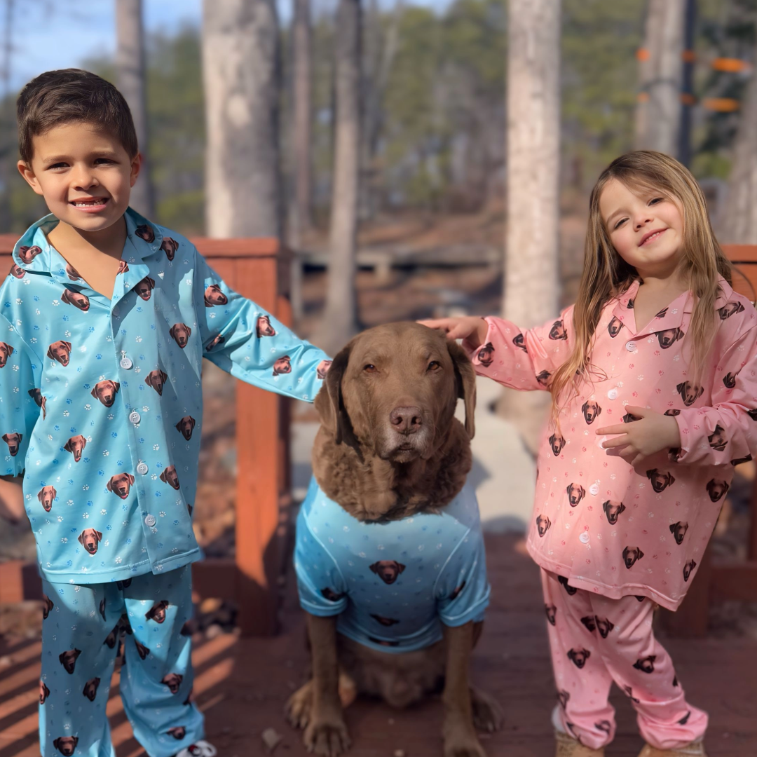 A young boy and girl stand on a wooden deck outdoors, each wearing custom pajamas printed with the face of their brown dog. The boy wears blue pajamas, the girl wears pink pajamas, and the large brown dog sits calmly between them wearing a matching blue shirt.
