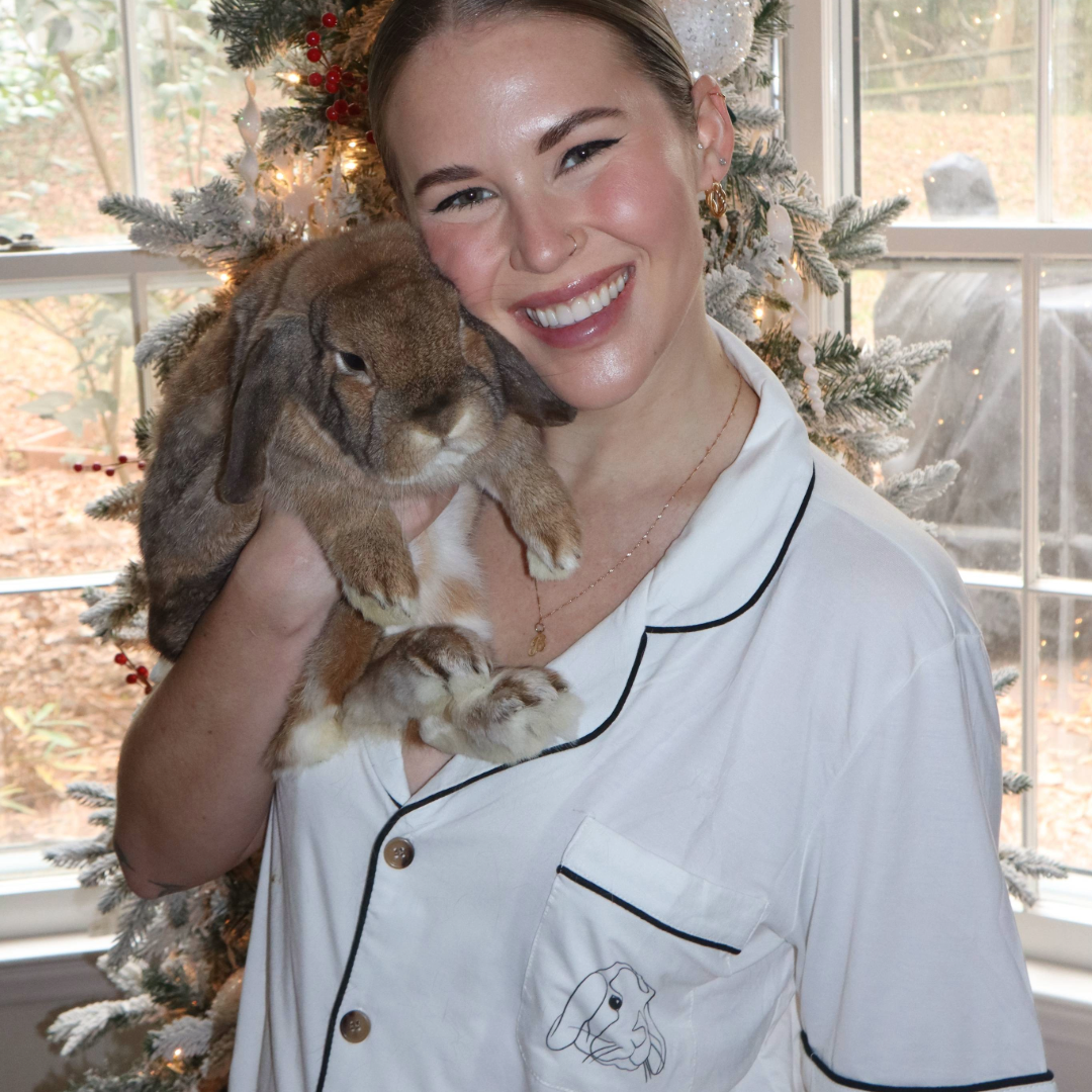 A woman wearing white pajamas with black trim and a small custom bunny illustration on the pocket smiles while holding a brown lop-eared rabbit against her cheek. They are sitting in front of a decorated, snow-flocked Christmas tree.