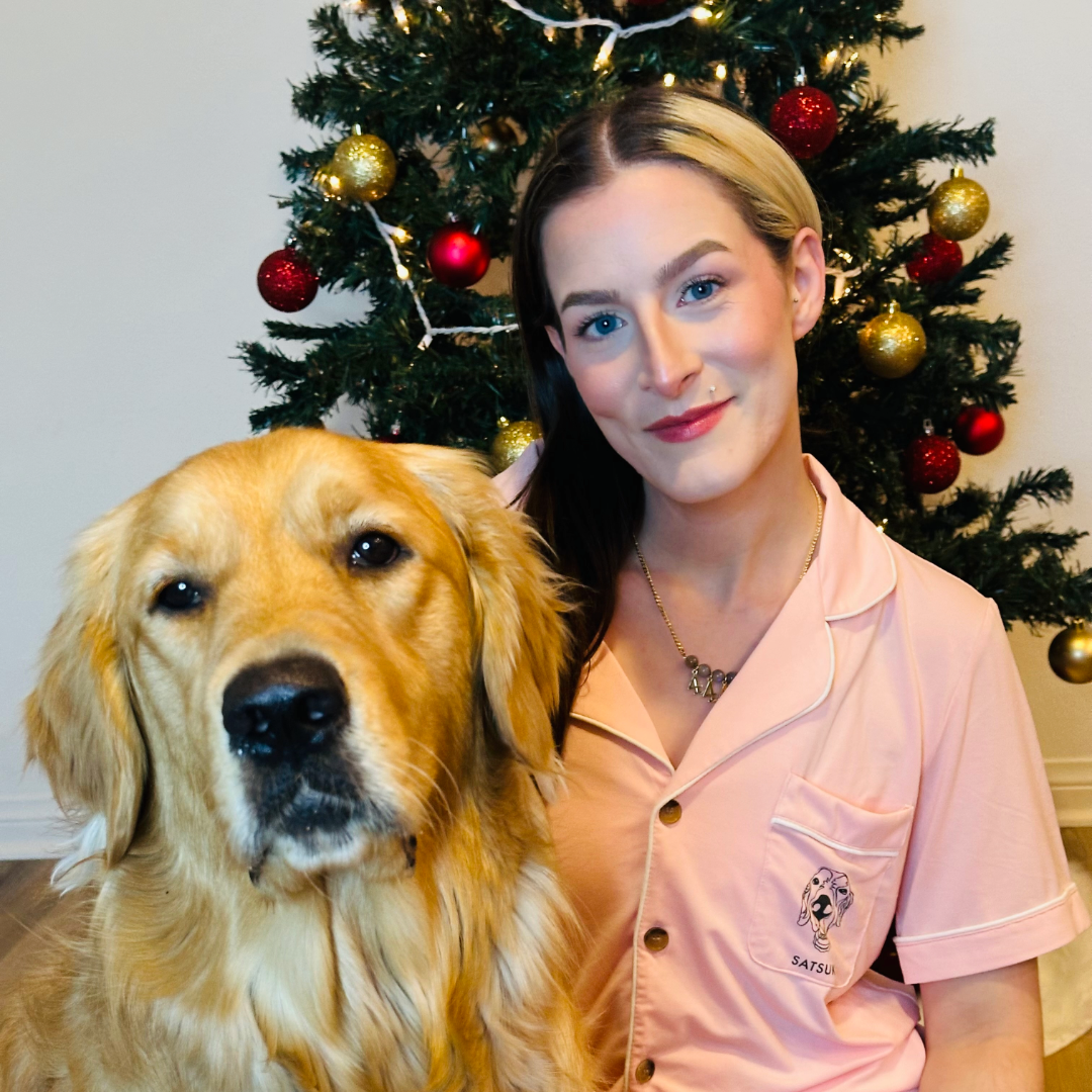 A woman in pink pajamas with a small custom dog illustration on the pocket sits beside a golden retriever in front of a decorated Christmas tree with red and gold ornaments.