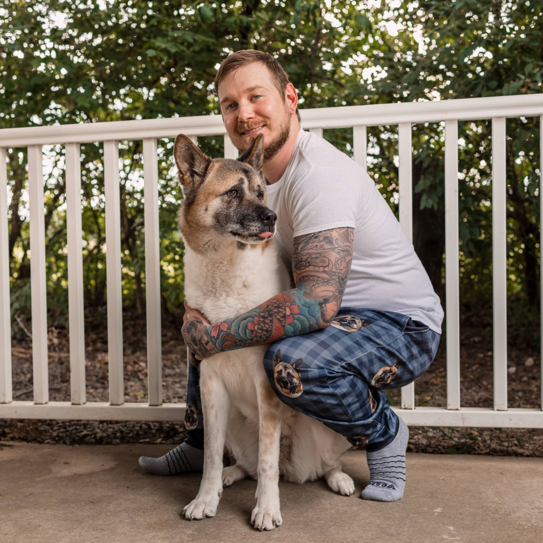 A man crouching on a porch, smiling as he hugs a large cream-and-brown dog. He is wearing blue plaid pajama pants printed with the dog’s face, along with a white T-shirt and patterned socks.
