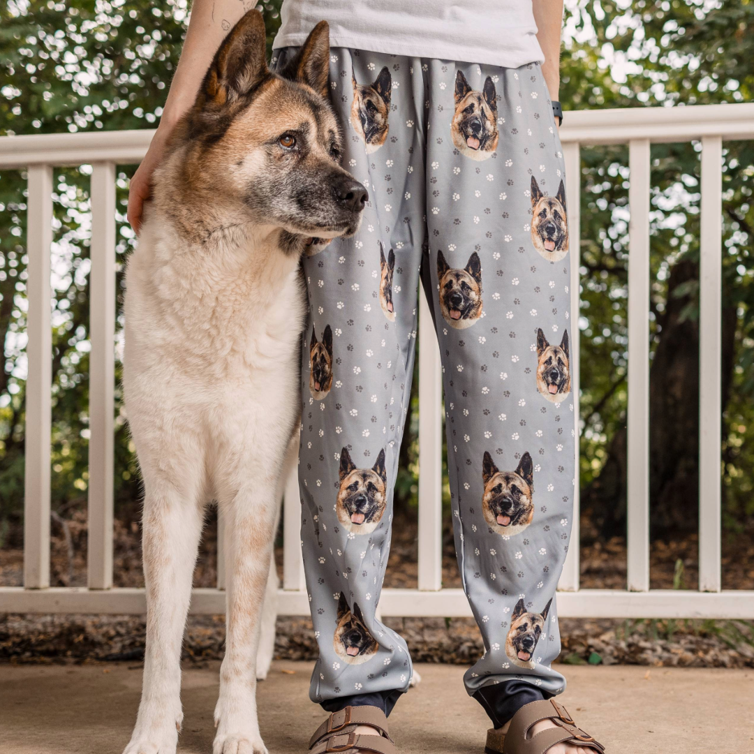 A large dog with a cream and brown coat stands beside a person wearing light gray pajama pants printed with the dog’s face and small white flower patterns. The person rests a hand on the dog’s back.