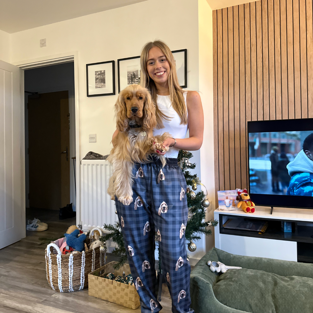 A woman standing in a living room holding a golden long-eared dog in her arms. She is smiling and wearing blue plaid pajama pants printed with the dog’s face.