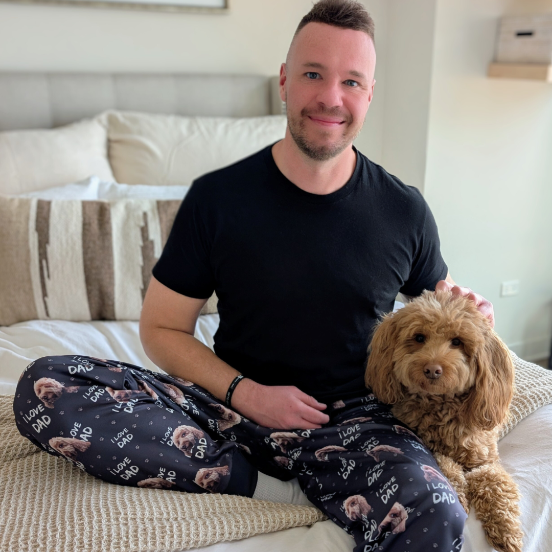 A man sitting cross-legged on a bed, wearing black “I Love Dad” pajama pants printed with a fluffy brown dog’s face. He is smiling and gently resting his hand on a matching brown, curly-haired dog sitting beside him.