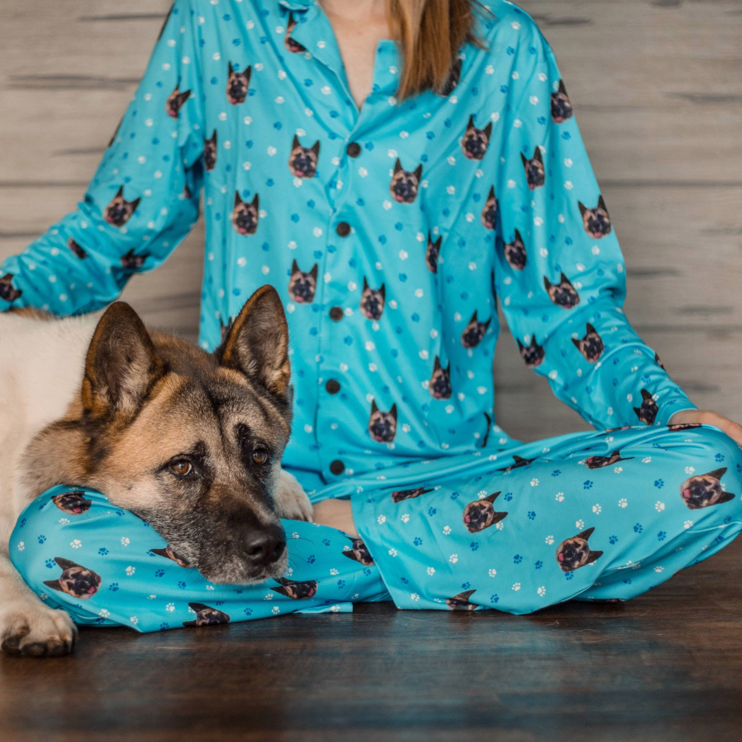 A person wearing bright blue pajamas printed with dog faces sits cross-legged on a wooden floor, while a large dog rests its head on their folded legs. The scene is cozy and calm, with the dog looking relaxed and content.
