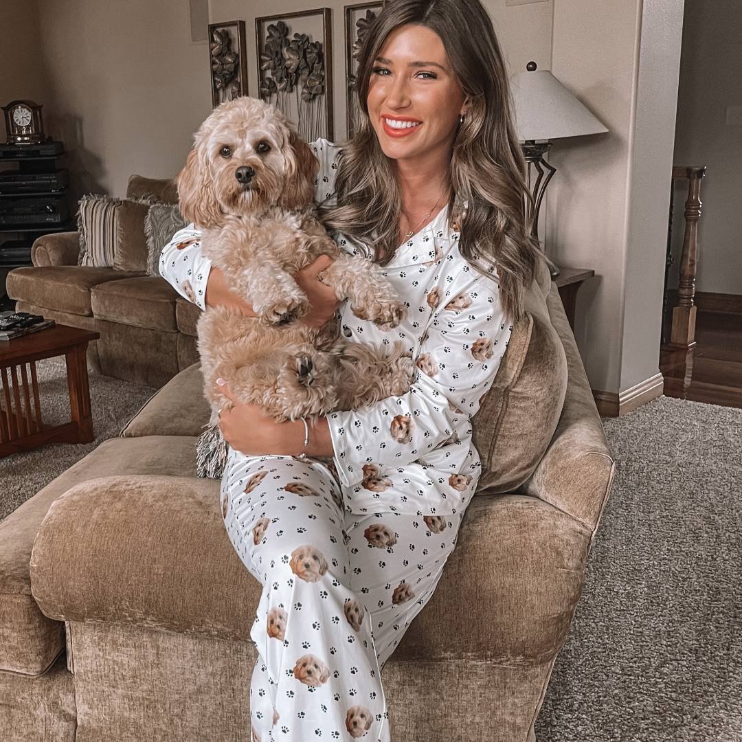 A smiling woman sitting on a beige couch wearing white pajamas printed with dog faces, holding a fluffy light-brown dog on her lap in a cozy living room.