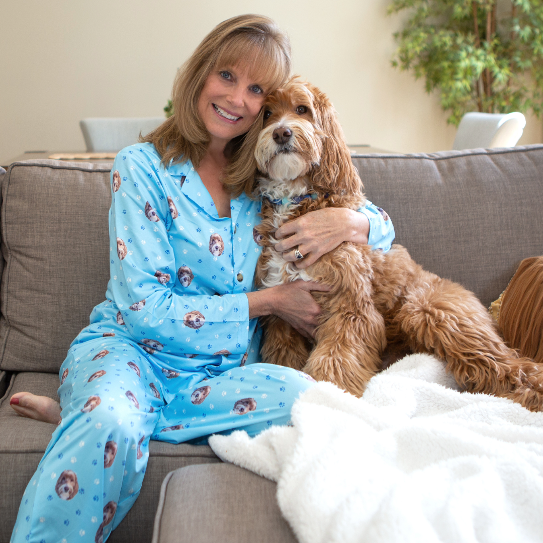 A woman sitting on a couch wearing light blue pajamas printed with dog faces, smiling as she hugs a fluffy brown and white dog resting beside her on a blanket.