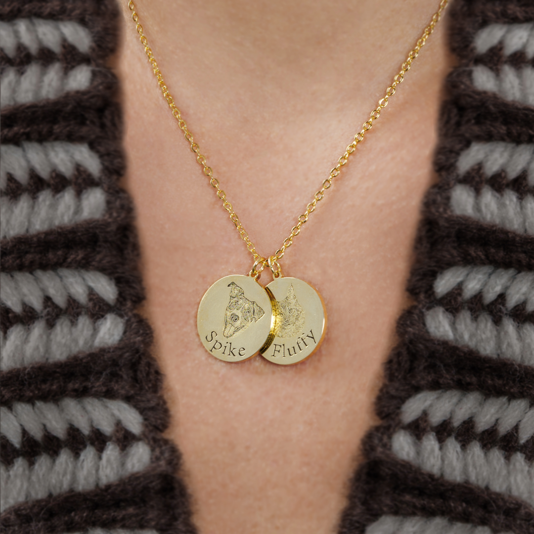 A close-up photo of a gold necklace worn around a person’s neck. The necklace has two round gold pendants engraved with pet portraits; one pendant says “Spike” and the other says “Fluffy.” The person is wearing a brown and gray knitted sweater.
