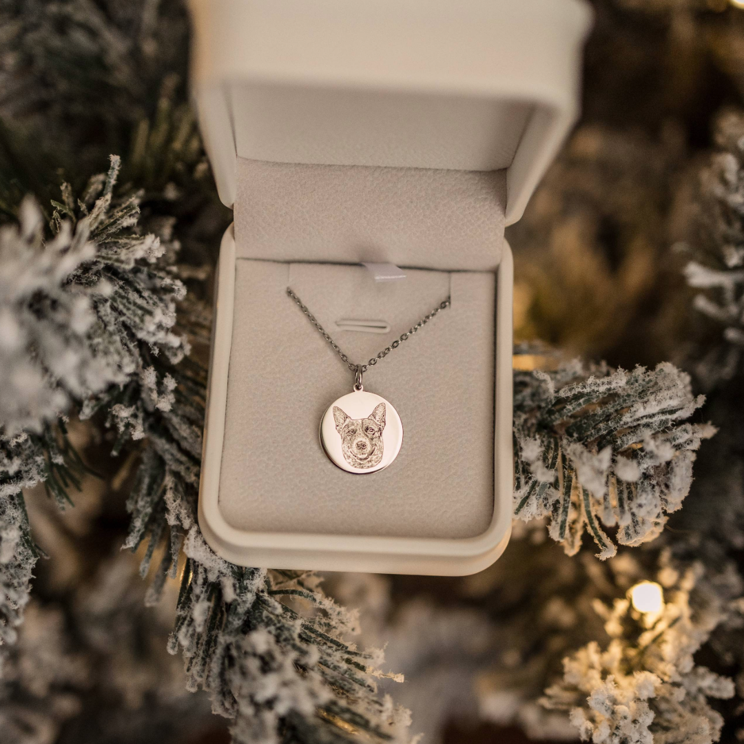 A silver necklace with a round pendant engraved with a dog’s face is displayed inside an open jewelry box. The box is set among snow-covered Christmas tree branches with warm lights glowing in the background.
