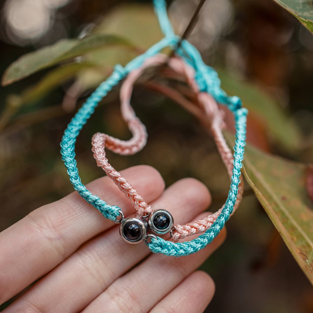 Two braided bracelets, one pink and one blue, with a black bead charm held between fingers against a blurred natural background.