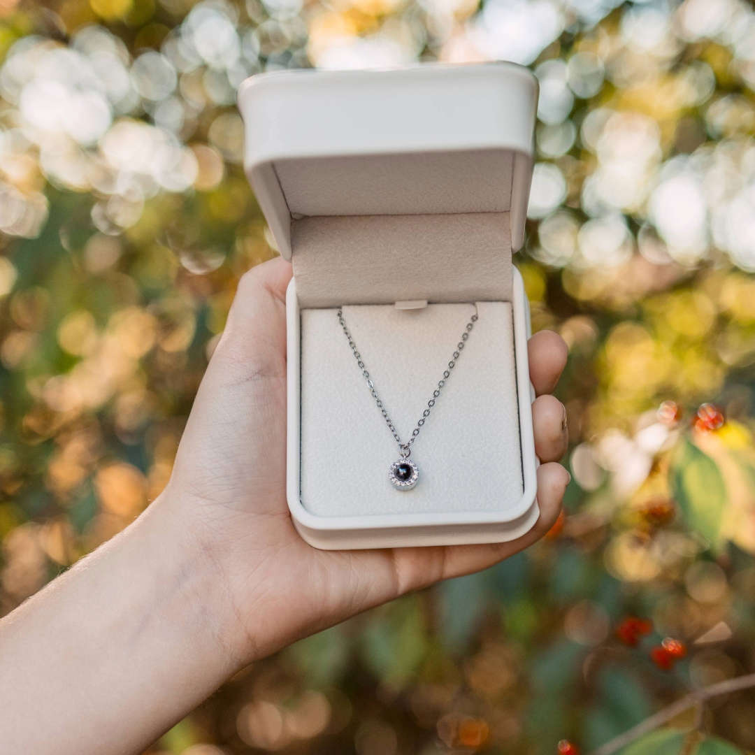 A close-up of a hand holding an open cream-colored jewelry box outdoors, with a silver necklace displayed inside. The background shows soft, warm bokeh from sunlit leaves and plants.