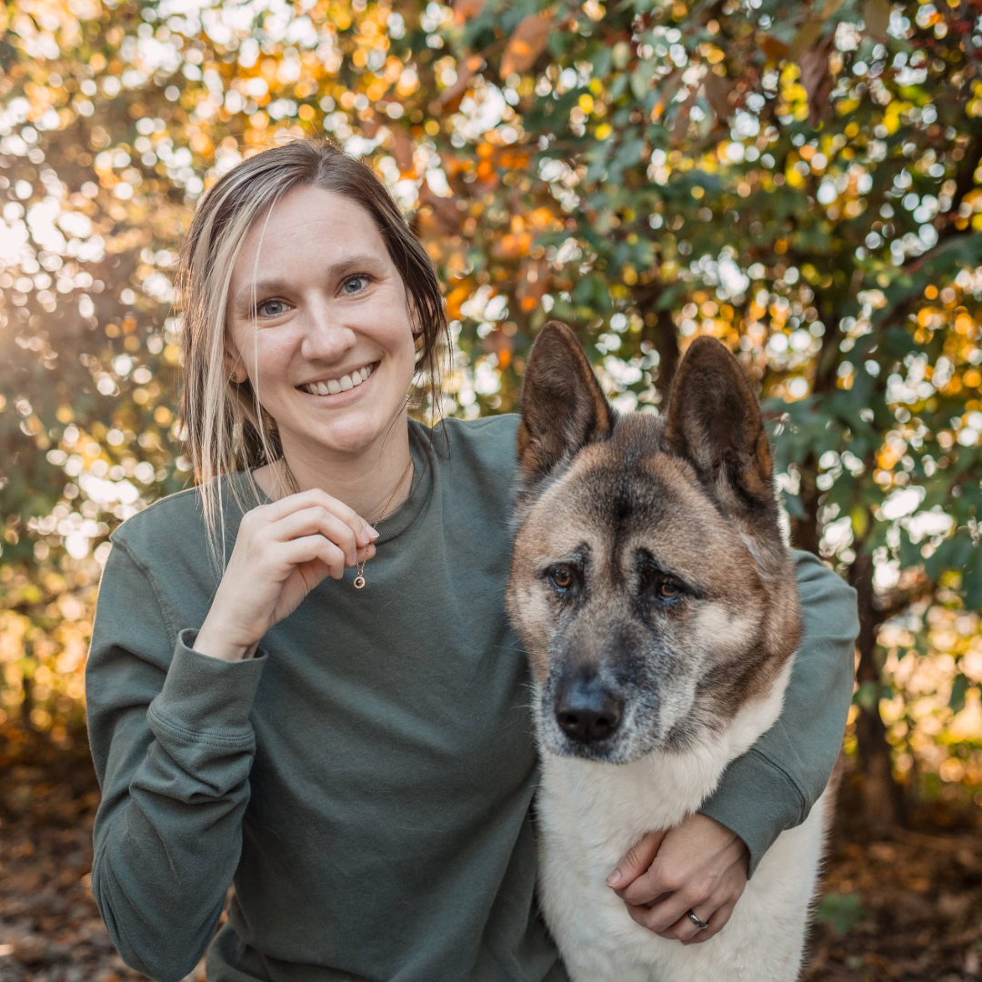 A woman outdoors smiles at the camera while holding a small necklace charm. She has one arm wrapped around a large Akita dog sitting beside her. Autumn leaves and soft sunlight fill the background.
