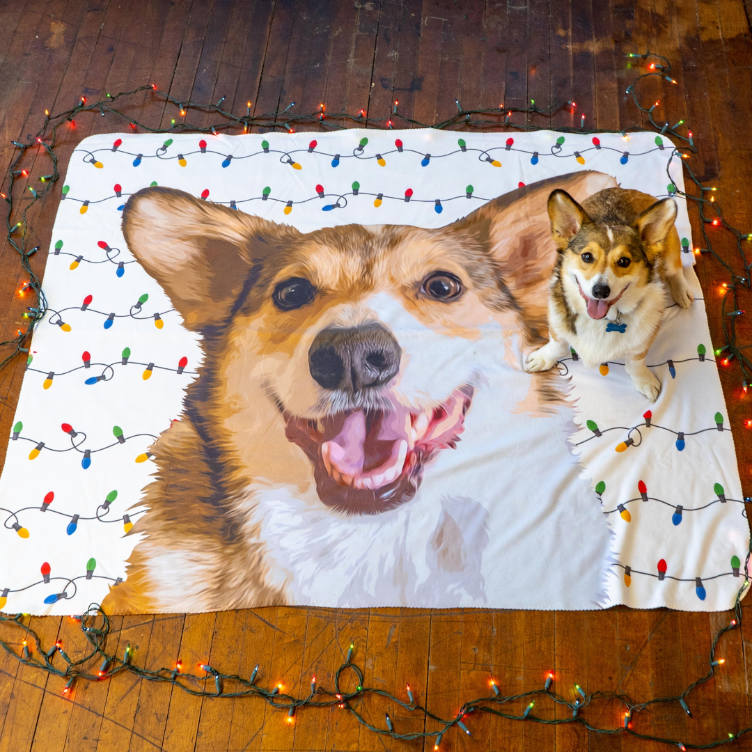 A Corgi sits on a wooden floor next to a large blanket printed with an illustrated portrait of the same dog. The blanket is decorated with colorful Christmas lights, with additional real string lights arranged around its edges.