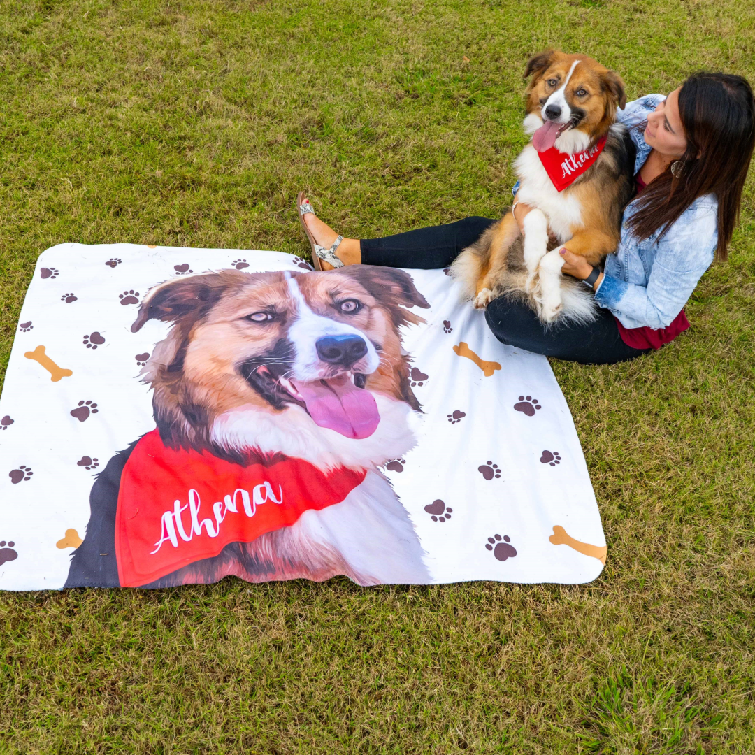 A woman sitting on the grass holds a brown and white dog wearing a red bandana that says “Athena.” In front of them is a white blanket printed with a large illustrated portrait of the same dog, also wearing the red bandana, along with paw prints and bone patterns.