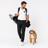 A man wearing a white polo shirt with a small bulldog graphic on the left chest, black trousers, and white sneakers walks while carrying a black golf bag with clubs over his shoulder. A matching bulldog is standing on the right against a clean white studio background.