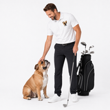 A man in a white polo shirt with a dog printed logo and black trousers stands next to a black golf bag and club, petting a brown and white bulldog on a white studio background.