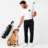 A man wearing a white polo shirt with a bulldog graphic on the chest, black trousers, and white sneakers stands next to a dog while holding a golf club. A golf bag is visible on the left against a clean white studio background.