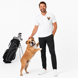 A man wearing a white polo shirt with a small bulldog graphic on the left chest stands next to a black golf bag and a brown bulldog on a white studio background.