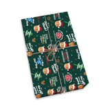 A gift-wrapped rectangular package photographed on a clean white background. The wrapping paper is dark green with a Christmas-themed print featuring Santa hats, cookies, ornaments, and phrases like 'A Christmas Dad' and 'The Toy.
