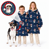 Two children and a dog wearing matching navy blue hooded blankets with a Christmas dog print, shown on a white studio background. One child has a protective face shield, and a circular inset shows a close-up of the printed design on the blanket.
