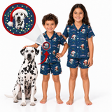 Two children wearing matching navy blue Christmas dog pajamas stand barefoot on a white studio background next to a Dalmatian. A circular inset at the top left shows a close-up of the pajama print with a dog wearing a Santa hat.
