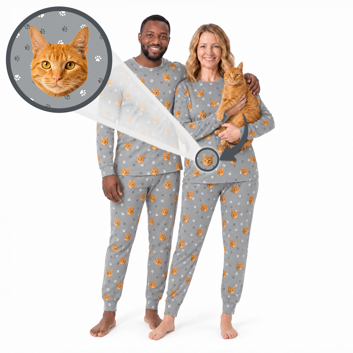 A man and a woman wearing matching gray hooded pajamas with an orange and white cat face print, standing barefoot on a white background while holding an orange cat. A circular inset shows a close-up of the cat face print on the pajama fabric.