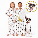 Two children wearing matching white pajamas with a dog print stand barefoot against a white background, with a circular inset showing a dog wearing the pajamas and a yellow arrow pointing to the child's pajama top.