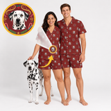 A man and a woman wearing matching burgundy short-sleeve pajama sets with a Dalmatian dog print, standing barefoot on a white studio background next to a Dalmatian dog. A circular inset at the top left shows a close-up of the printed dog motif, and a yellow arrow points from the inset to the pattern on the pajamas.