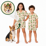 Two children wearing matching short-sleeve pajama sets with a cactus and Santa Claus gift print, standing barefoot on a white background next to a corgi. An inset circle at the top left shows a custom dog portrait with a Santa hat, and arrows point to matching pajama prints on the children's clothing.
