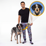 A man and a blue merle dog stand on a white studio background wearing matching custom pet apparel. The man wears gray patterned sweatpants with a dog graphic, and the dog wears matching dog-print socks.