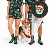 A couple wearing matching dark green Christmas-themed pajama sets and socks stands with a corgi dog on a white studio background. The pajamas and socks have a festive print with text like 'Merry Christmas' and holiday graphics.
