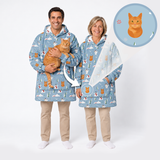 Two adults wearing matching light blue hooded blankets with a playful cat print, standing against a white studio background. One man holds an orange tabby cat, and a circular inset shows a close-up of the cat motif on the blanket.