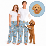 A smiling couple wearing matching light blue custom pet-print pajama pants stands beside their curly-haired dog. A magnified callout highlights the dog’s illustrated face printed throughout the personalized pajama pattern.