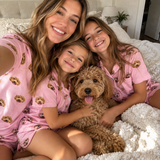 A family shot of a mom and her two daughters wearing matching pink pajamas with a brown dog lying on a white fluffy bed. The pajamas feature a repeating pattern of a brown and cream dog face.