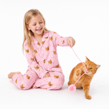 Child wearing a Light Pink Paws cat-print pajama set sitting on the floor and playing with an orange cat using a pink toy.