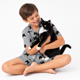 Child wearing a slate gray cat-print pajama set sitting cross-legged and holding a black and white cat.