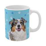 Close up of light blue ceramic mug with paws pattern, featuring a smiling, multicolored Australian Shepherd