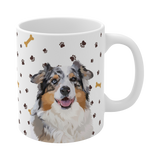 Close up of white ceramic mug with paws and bones pattern, featuring a smiling, multicolored Australian Shepherd