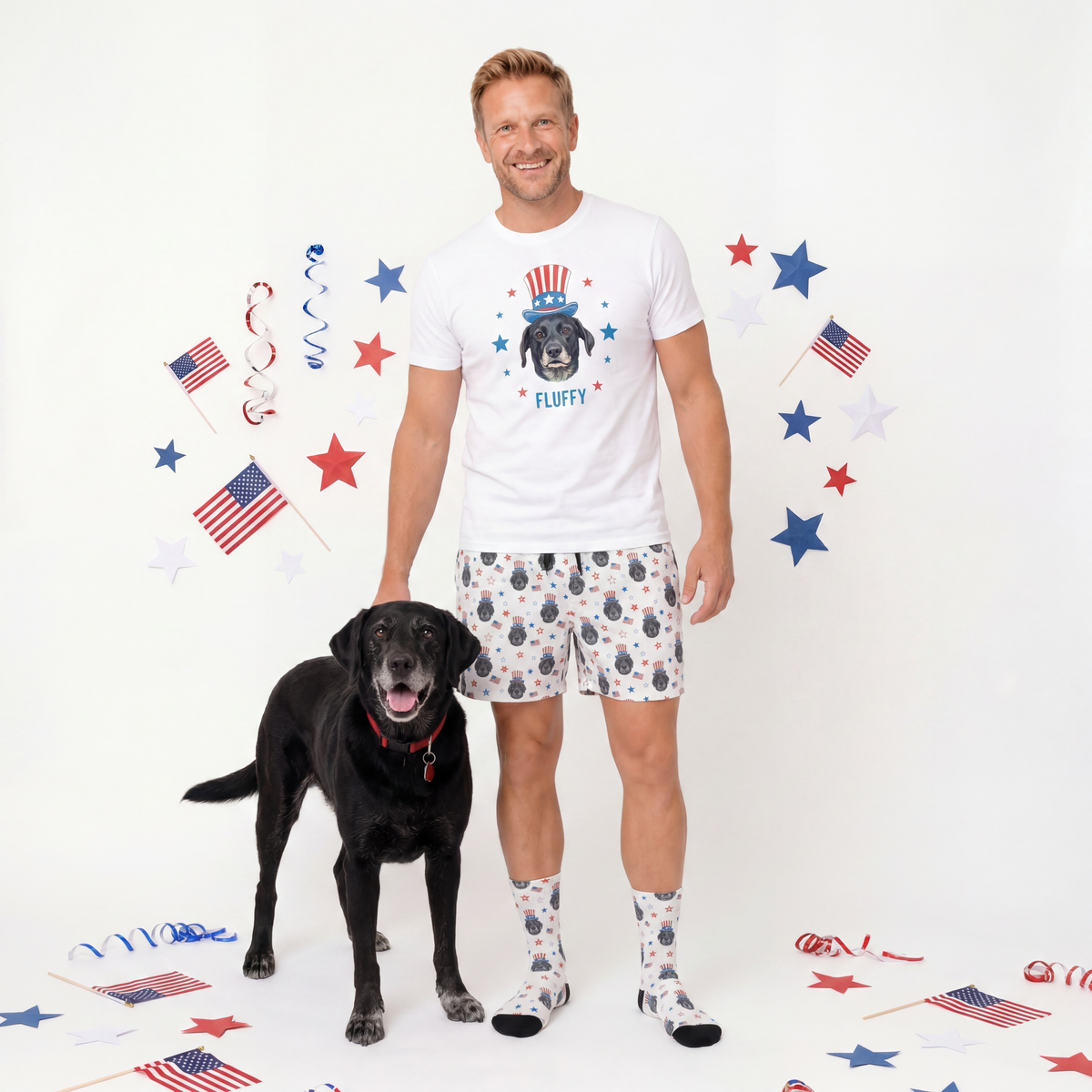 Man wearing a white t-shirt with a dog graphic and patterned shorts, standing next to a black dog on a white background with patriotic decorations.