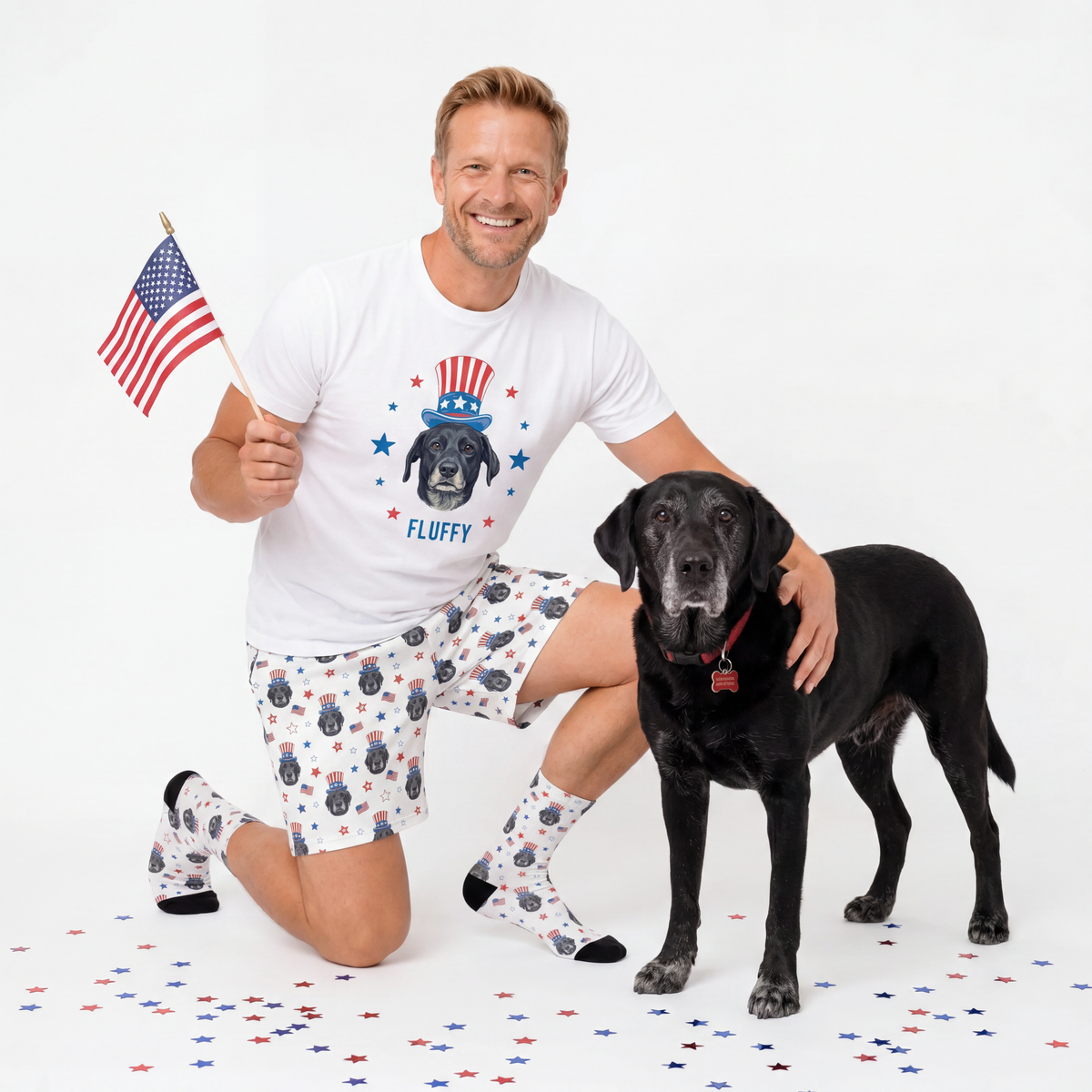 Man wearing a white t-shirt with a dog graphic and text, holding an American flag, and kneeling next to a black dog on a white background.