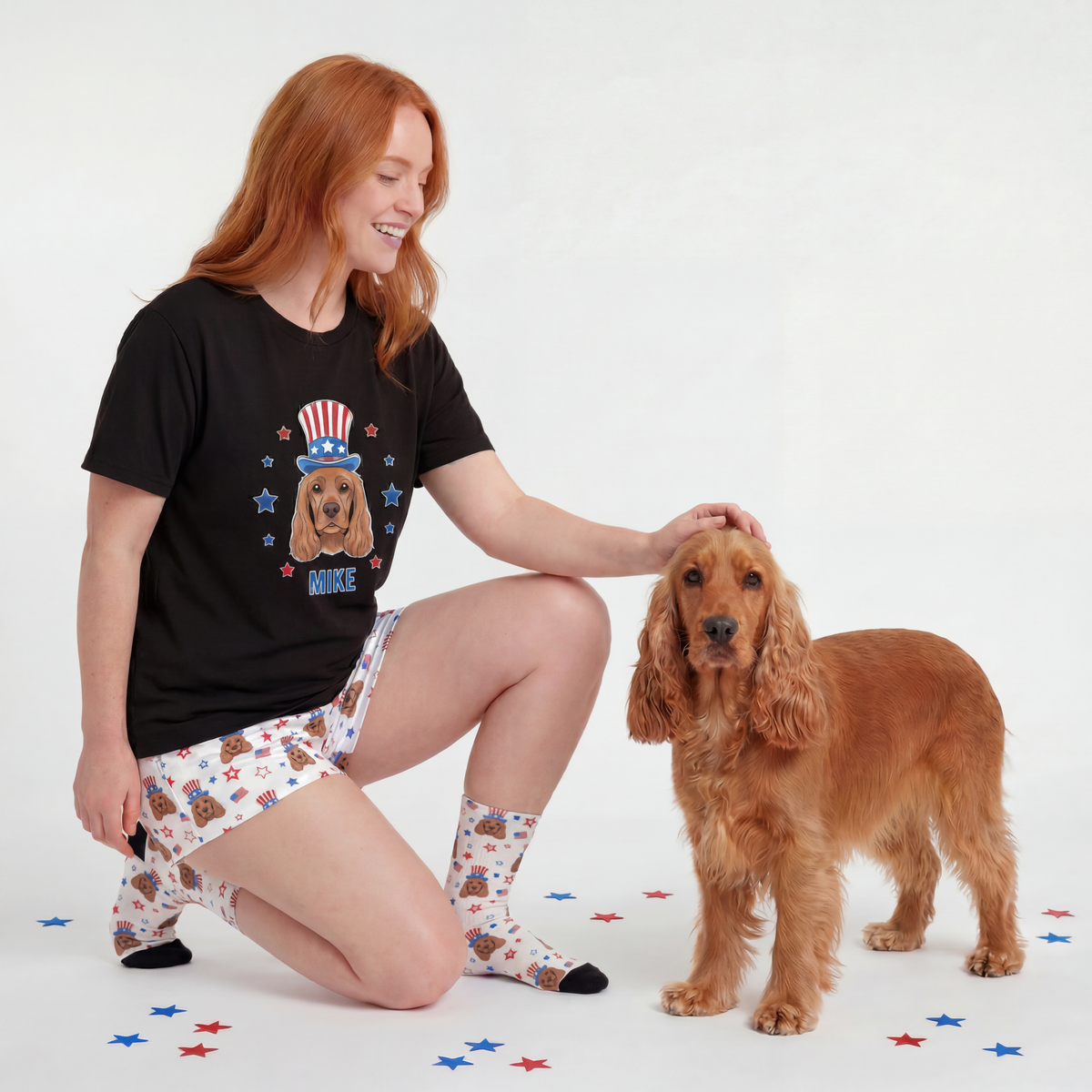 Woman wearing a black t-shirt with a dog graphic, white shorts with dog pattern, and socks with dog pattern, kneeling next to a brown dog on a white background.