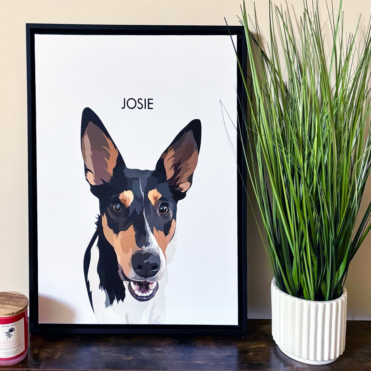 Framed custom pet portrait featuring an illustrated black, tan, and white dog with large upright ears, displayed on a wooden surface. The name ‘Josie’ is printed above the portrait, with a potted green plant and a candle placed beside the frame.